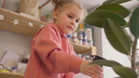 Bottom-View-Of-A-Little-Girl-Spraying-Water-On-A-Plant-At-A-Table-In-A-Craft-Workshop