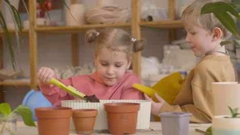 Little-Blonde-Girl-And-Blond-Kid-Preparing-The-Soil-A-Watering-A-Pot-Sitting-At-A-Table-Where-Is-Plants-In-A-Craft-Workshop