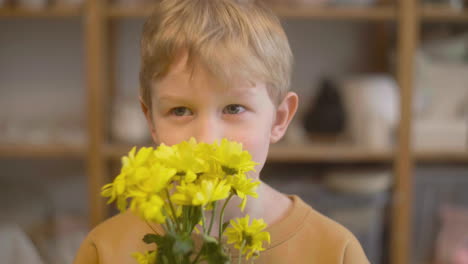 Close-Up-View-Of-A-Blond-Kid-Smelling-Yellow-Flowers-And-Looking-At-Camera-In-A-Craft-Workshop