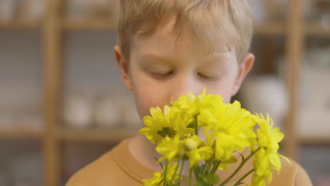Close-Up-View-Of-A-Blond-Kid-Smelling-Yellow-Flowers-And-Looking-At-Camera-In-A-Craft-Workshop-1