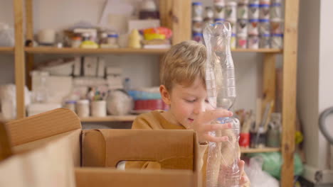 Blond-Kid-Putting-Plastic-Bottles-In-Cardboard-Box-On-A-Table-In-A-Craft-Workshop