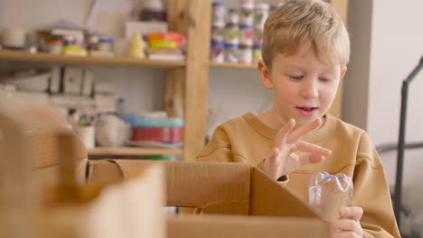 Blond-Kid-Looking-A-Plastic-Bottle-And-Putting-It-In-Cardboard-Box-On-A-Table-In-A-Craft-Workshop