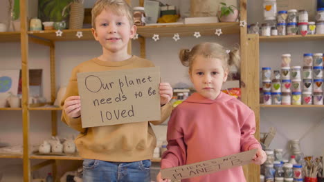 Blond-Boy-And-Blonde-Little-Girl-In-Craft-Workshop-Holding-Two-Cardboard-Signs-With-There's-No-Planet-B-And-Our-Planet-Needs-To-Be-Loved-Phrases