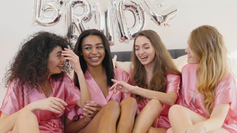 Group-Of-Multiethnic-Female-Friends-Dressed-In-Pink-Silk-Nightdresses-Talking-And-Sitting-On-Bed-Decorated-With-Word-'Bride'-Ballons-In-Bridal-Gathering-1