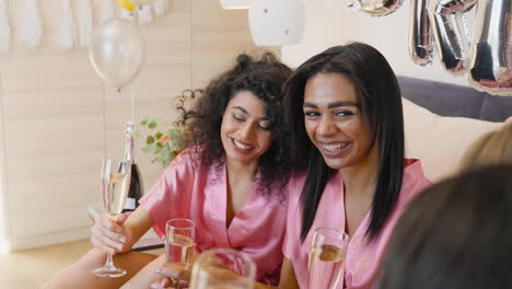 Eine-Gruppe-Multiethnischer-Freundinnen-In-Rosa-Seidennachthemden,-Die-Sich-Unterhalten,-Während-Sie-Mit-Champagnergläsern-Anstoßen