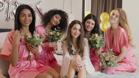 Group-Of-Multiethnic-Female-Friends-And-Bride-Who-Is-Wearing-White-Silk-Nightdresses-Looking-At-Camera-Sitting-On-Bed-Holding-Bouquets-Decorated-With-Word-'Bride'-Ballons-In-Bridal-Gathering