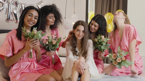 Group-Of-Multiethnic-Female-Friends-And-Bride-Who-Is-Wearing-White-Silk-Nightdresses-Looking-At-Camera-Sitting-On-Bed-Holding-Bouquets-Decorated-With-Word-'Bride'-Ballons-In-Bridal-Gathering-1