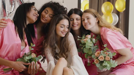 Group-Of-Multiethnic-Female-Friends-Caressing-Bride-Who-Is-Wearing-White-Silk-Nightdresses-Sitting-On-Bed-Holding-Bouquets-Decorated-With-Word-'Bride'-Ballons-In-Bridal-Gathering