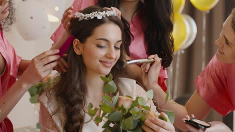 Group-Of-Female-Friends-Making-Up-The-Bride-And-Putting-Her-A-Hair-Band-Holding-A-Bouquet-Sitting-On-Bed-In-Bridal-Gathering-1