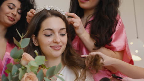 Close-Up-View-Of-Group-Of-Female-Friends-Making-Up-The-Bride-And-Putting-Her-A-Hair-Band-While-Holding-A-Bouquet-Sitting-On-Bed-In-Bridal-Gathering
