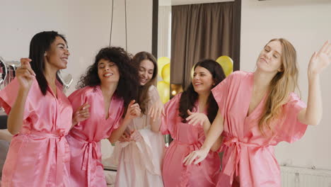 Gruppe-Multiethnischer-Freundinnen-Und-Braut,-Die-Rosafarbene-Und-Weiße-Seidennachthemden-Tragen,-Während-Sie-In-Einem-Raum-In-Der-Brautversammlung-Tanzen