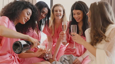 Gruppe-Multiethnischer-Freundinnen-Und-Braut-Tragen-Rosa-Und-Weiße-Seidennachtkleider,-Während-Sie-Champagnergläser-In-Einem-Raum-In-Der-Brautversammlung-Halten