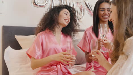 Group-Of-Multiethnic-Female-Friends-And-Bride-Wearing-Pink-And-White-Silk-Nighdresses-While-Toasting-With-Champagne-Glasses-In-A-Room-In-Bridal-Gathering