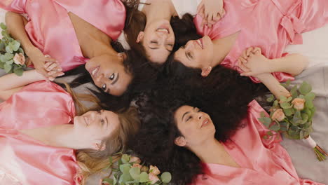 Top-View-Of-Group-Of-Multiethnic-Female-Friends-And-Bride-Dressed-In-Pink-And-White-Silk-Nightdresses-Holding-Hands-With-Bouquets-And-Laughing-While-Laying-On-Floor