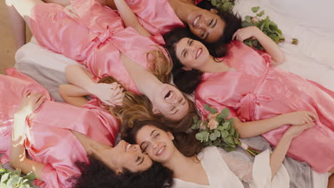 Blick-Von-Oben-Auf-Eine-Gruppe-Multiethnischer-Freundinnen-Und-Braut-In-Rosa-Und-Weißen-Nachthemden-Aus-Seide,-Die-Händchen-Mit-Blumensträußen-Halten-Und-Lachen,-Während-Sie-Auf-Etage-1-Liegen