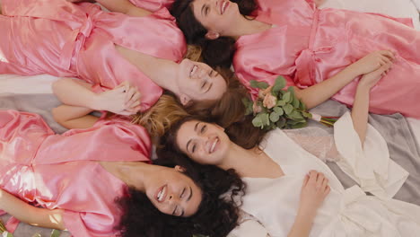 Top-View-Of-Group-Of-Multiethnic-Female-Friends-And-Bride-Dressed-In-Pink-And-White-Silk-Nightdresses-Holding-Hands-With-Bouquets-And-Laughing-While-Laying-On-Floor-2