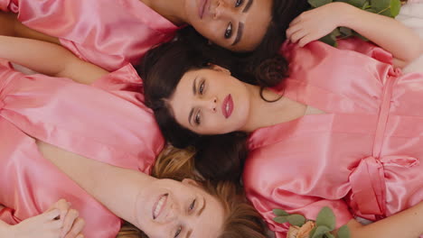 Top-View-Of-Group-Of-Multiethnic-Female-Friends-And-Bride-Dressed-In-Pink-And-White-Silk-Nightdresses-Holding-Hands-With-Bouquets-While-Laying-On-Floor