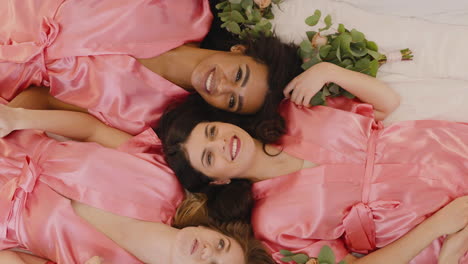 Blick-Von-Oben-Auf-Eine-Gruppe-Multiethnischer-Freundinnen-Und-Braut-In-Rosa-Und-Weißen-Nachthemden-Aus-Seide,-Die-Händchen-Mit-Blumensträußen-Halten-Und-Lachen,-Während-Sie-Auf-Etage-3-Liegen