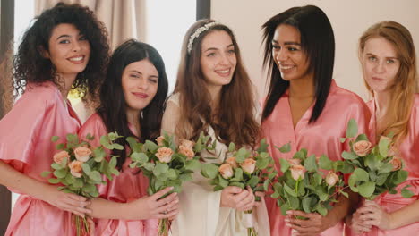 Group-Of-Multiethnic-Female-Friends-And-Bride-Looking-At-Camera,-Wearing-Silk-Pink-And-White-Nighdresses-While-Holding-Bouquets