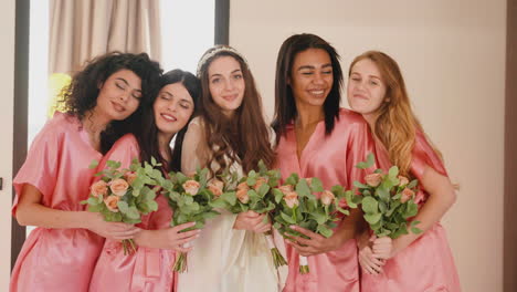 Gruppe-Multiethnischer-Freundinnen-Und-Braut,-Die-In-Die-Kamera-Schauen-Und-Seidenrosa-Und-Weiße-Nachthemden-Tragen,-Während-Sie-Blumensträuße-Halten-1