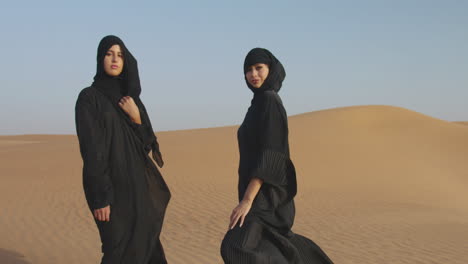 Dos-Hermosas-Mujeres-Musulmanas-En-Hiyab-Posando-En-Un-Desierto-Ventoso-Y-Mirando-A-La-Cámara
