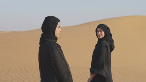 Dos-Hermosas-Mujeres-Musulmanas-En-Hiyab-De-Pie-En-Un-Desierto-Ventoso-Y-Sonriendo-A-La-Cámara