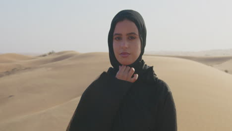 Porträt-Einer-Schönen-Muslimischen-Frau-Mit-Hijab,-Die-In-Einer-Windigen-Wüste-In-Die-Kamera-Blickt