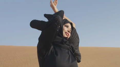 Muslimische-Frau-Mit-Traditionellem-Schwarzem-Kleid-Und-Hijab,-Die-In-Einer-Windigen-Wüste-Posiert