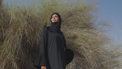 Schöne-Muslimische-Frau-In-Traditioneller-Kleidung-Und-Hijab-Posiert-In-Der-Nähe-Eines-Wüstenstrauchs