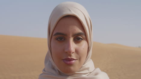 Nahaufnahme-Des-Porträts-Einer-Schönen-Muslimischen-Frau-Im-Hijab,-Die-In-Einer-Windigen-Wüste-Steht-Und-In-Die-Kamera-Blickt