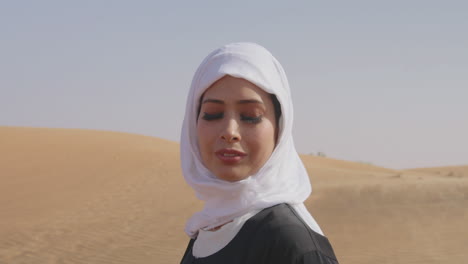 Porträt-Einer-Schönen-Muslimischen-Frau-In-Weißem-Hijab-Und-Traditionellem-Schwarzem-Kleid,-Die-In-Einer-Windigen-Wüste-Steht-Und-In-Die-Kamera-Lächelt