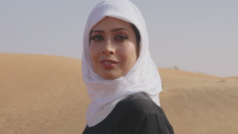 Porträt-Einer-Schönen-Muslimischen-Frau-In-Weißem-Hijab-Und-Traditionellem-Schwarzem-Kleid,-Die-In-Einer-Windigen-Wüste-Steht-Und-In-Die-Kamera-2-Lächelt