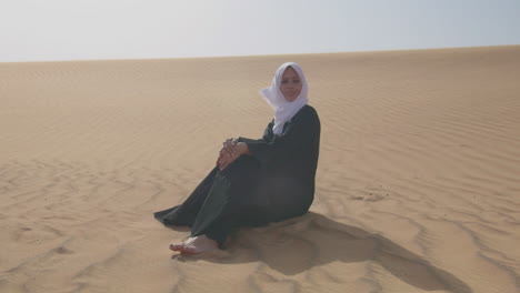 Beautiful-Muslim-Woman-In-White-Hijab-And-Traditional-Black-Dress-Sitting-On-Sand-In-A-Windy-Desert
