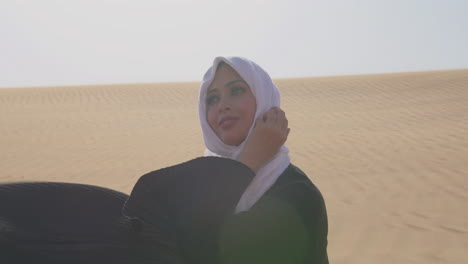 Mujer-Musulmana-Con-Hiyab-Blanco-Y-Vestido-Negro-Tradicional-Sentada-En-La-Arena-En-Un-Desierto-Ventoso-Y-Sonriendo-A-La-Cámara