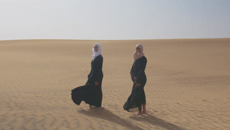 Two-Muslim-Women-Wearing-Traditional-Dress-And-Hijab-Standing-In-A-Windy-Desert-And-Looking-At-Camera