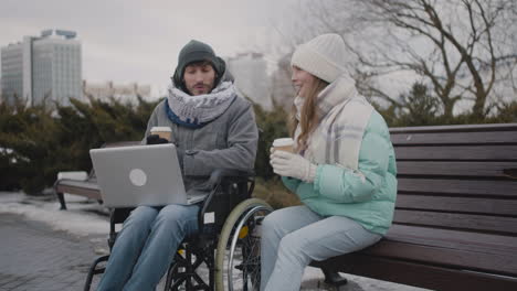Behinderter-Mann-Im-Rollstuhl-Und-Sein-Freund-Sehen-Sich-Etwas-Lustiges-Auf-Dem-Laptop-An,-Während-Sie-Im-Winter-1-Im-Stadtpark-Kaffee-Zum-Mitnehmen-Trinken