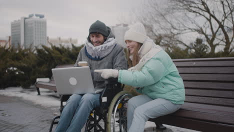 Behinderter-Mann-Im-Rollstuhl-Und-Sein-Freund-Schauen-Sich-Etwas-Lustiges-Auf-Dem-Laptop-An,-Während-Sie-Im-Winter-2-Im-Städtischen-Park-Kaffee-Zum-Mitnehmen-Trinken