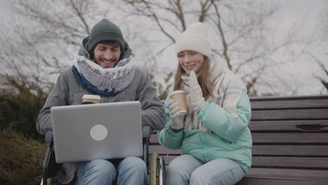 Disabled-Man-In-Wheelchair-And-His-Friend-Watching-Something-Funny-On-Laptop-Computer-And-Laughing-Together-At-Urban-Park-In-Winter