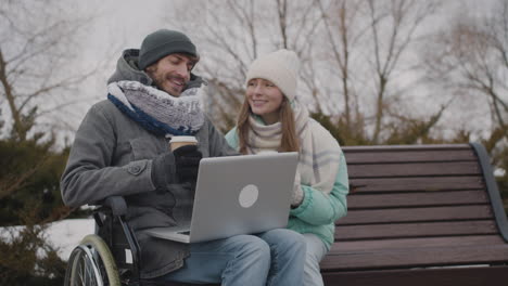 Disabled-Man-In-Wheelchair-And-His-Friend-Watching-Something-Funny-On-Laptop-Computer-While-Drinking-Takeaway-Coffee-At-Urban-Park-In-Winter-4