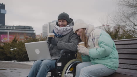 Behinderter-Mann-Im-Rollstuhl-Und-Sein-Freund-Schauen-Sich-Etwas-Lustiges-Auf-Dem-Laptop-An-Und-Lachen-Zusammen-Im-Stadtpark-Im-Winter-1