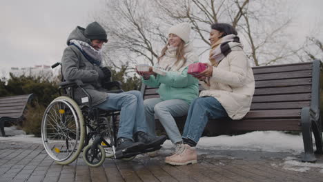 Two-Women-With-Small-Cake-And-A-Gift-Singing-Happy-Birthday-To-Their-Disabled-Friend-At-Urban-Park-In-Winter