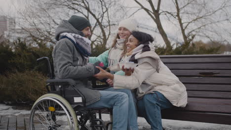 Two-Women-With-Small-Birthday-Cake-And-A-Gift-Giving-A-Surprise-To-Their-Disabled-Friend-In-Wheelchair-At-Urban-Park-In-Winter