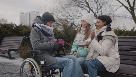 Happy-Disabled-Man-In-Wheelchair-Opening-A-Gift-Box-While-Celebrating-His-Birthday-With-Two-Friends-At-Urban-Park-In-Winter