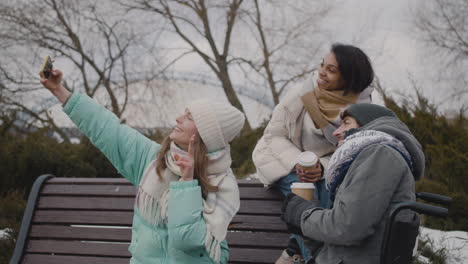 Grupo-De-Amigos,-Dos-Mujeres-Y-Un-Hombre-Discapacitado-En-Silla-De-Ruedas,-Tomando-Un-Video-Selfie-En-Un-Parque-Urbano-Durante-La-Temporada-De-Invierno