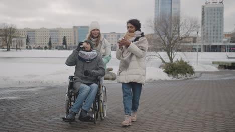 Behinderter-Mann-Im-Rollstuhl-Und-Zwei-Frauen,-Die-Zusammen-Gehen-Und-Im-Winter-Kaffee-Zum-Mitnehmen-In-Der-Stadt-Trinken