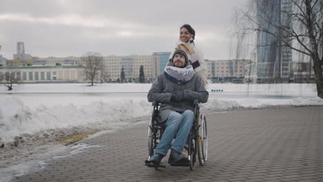 Fröhliche-Muslimische-Frau,-Die-Im-Winter-Mit-Ihrem-Behinderten-Freund-Im-Rollstuhl-Durch-Die-Stadt-Spaziert