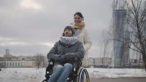 Glückliche-Muslimische-Frau,-Die-Im-Winter-1-Ihren-Behinderten-Freund-Im-Rollstuhl-Auf-Einem-Spaziergang-Durch-Die-Stadt-Mitnimmt