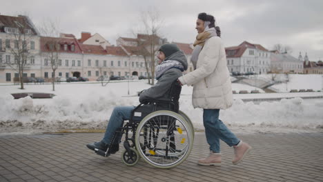 Seitenansicht-Einer-Glücklichen-Muslimischen-Frau,-Die-Im-Winter-Mit-Ihrem-Behinderten-Freund-Im-Rollstuhl-Durch-Die-Stadt-Spaziert