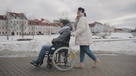Seitenansicht-Einer-Glücklichen-Muslimischen-Frau,-Die-Ihren-Behinderten-Freund-Im-Rollstuhl-Im-Winter-1-Zu-Einem-Spaziergang-Durch-Die-Stadt-Mitnimmt