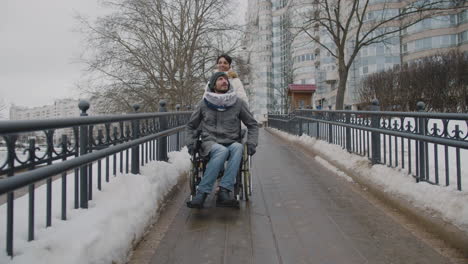 Vorderansicht-Einer-Muslimischen-Frau,-Die-Ihren-Behinderten-Freund-Im-Rollstuhl-Im-Winter-1-Auf-Einem-Spaziergang-In-Der-Stadt-Mitnimmt